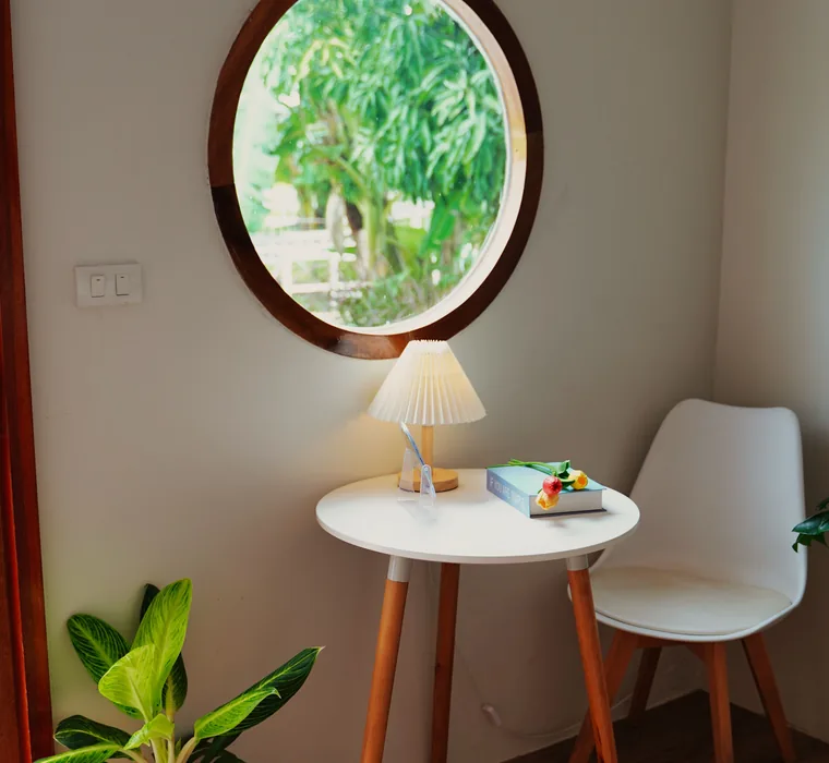 Góc phòng trọ có cửa sổ tròn, bàn nhỏ, đèn ngủ và chậu cây.